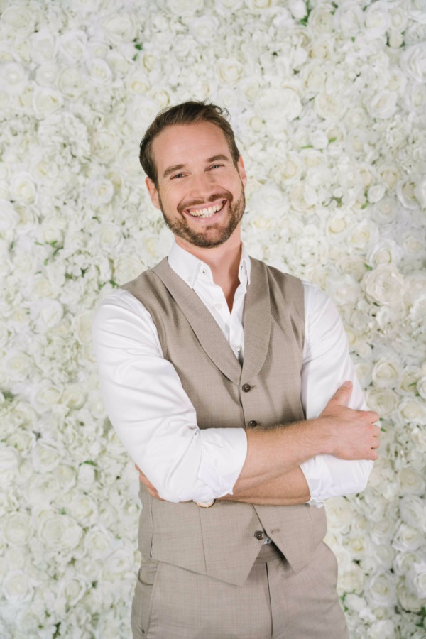Bräutigam lachend vor Blumenwand Pearl White Rosengold